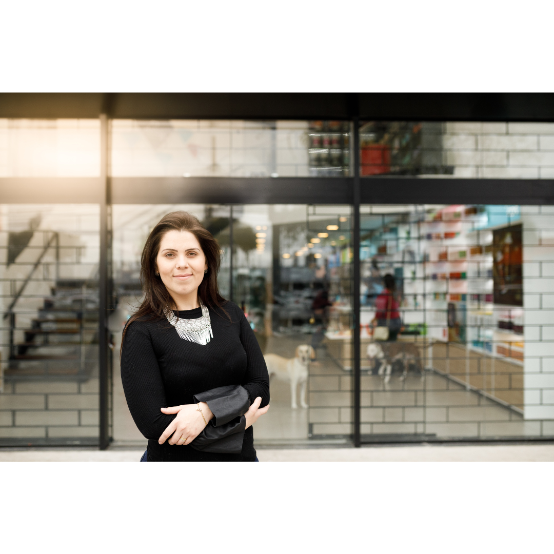 A confident woman stands outside a modern pet store with large glass windows, reflecting the bustling activity inside with customers and dogs.