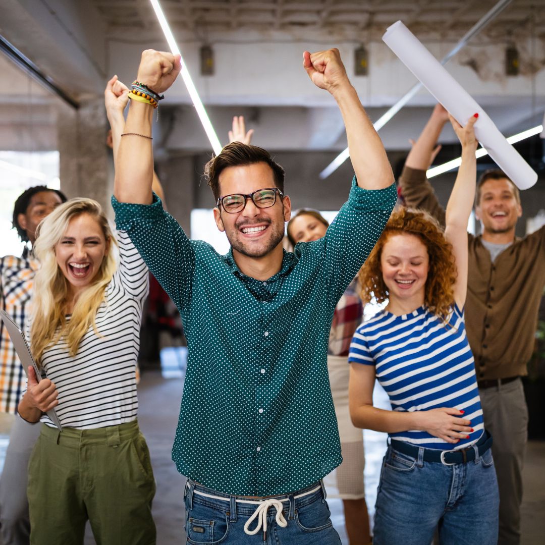 A cheerful group of young adults celebrate together indoors, smiling with raised arms.