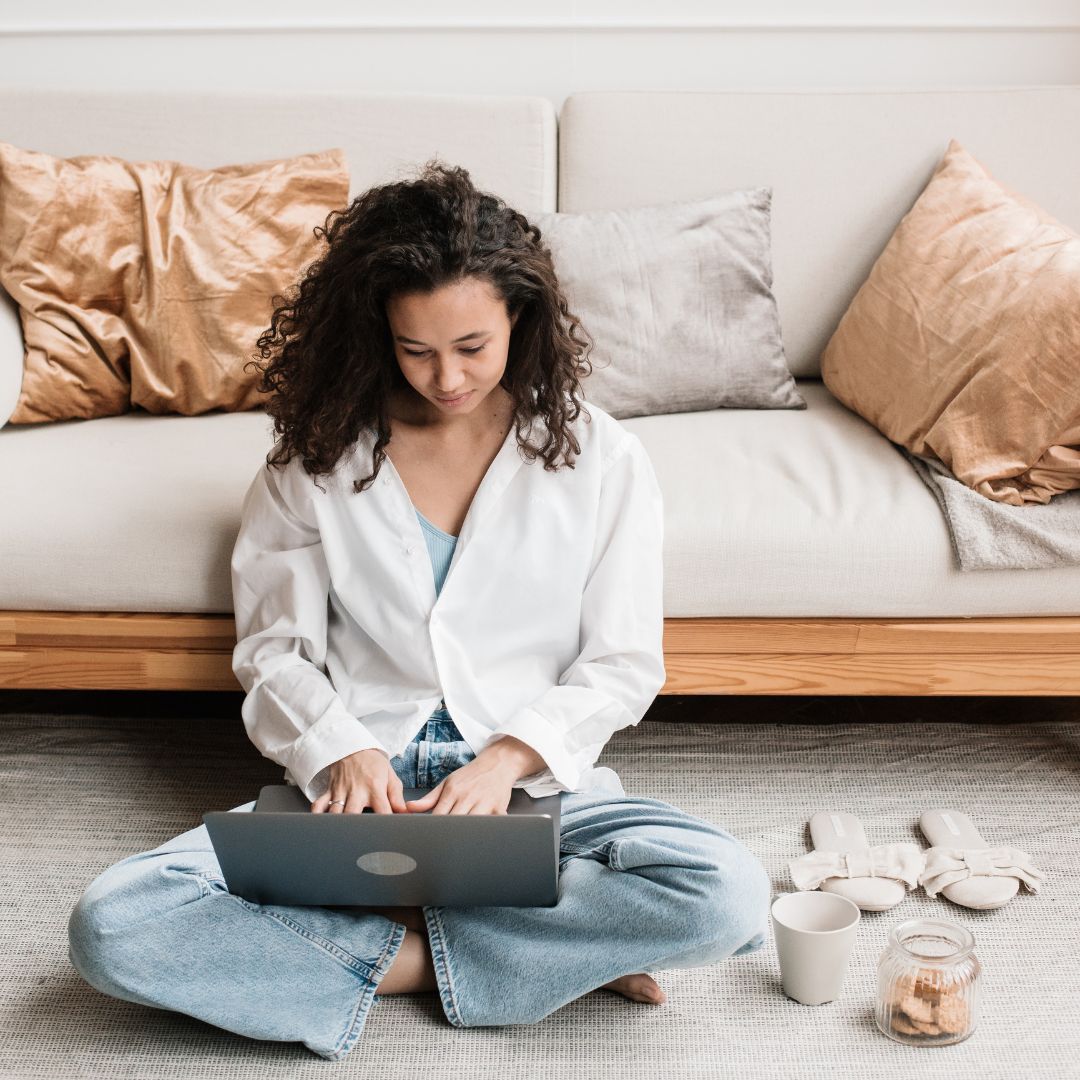 A young woman with curly hair sitting cross-legged on the floor, using a laptop in a cosy living room. She is wearing a white shirt and jeans, surrounded by neutral-coloured pillows and a small jar of cookies.