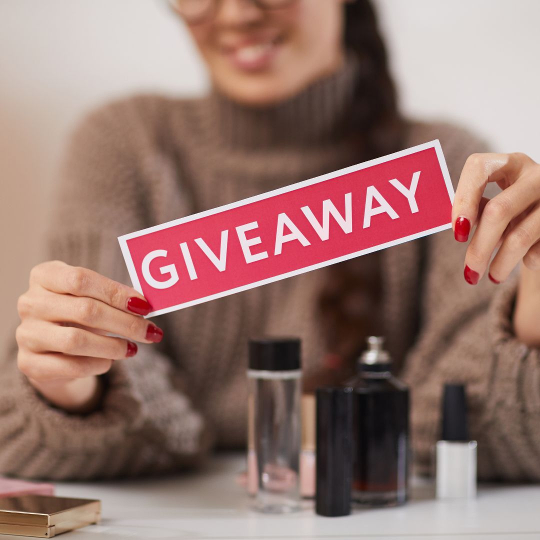 A woman with red-painted nails holding up a red sign with the word "GIVEAWAY" in bold white letters. She is smiling, and beauty products such as perfume bottles and nail polish are visible in front of her.