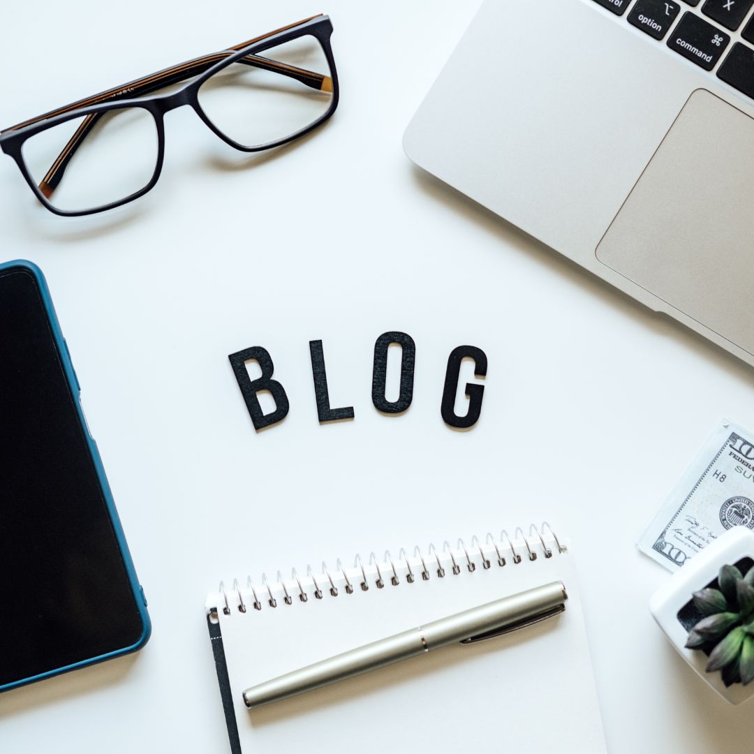 A flat lay of blogging essentials, including a laptop, glasses, a notepad, a pen, a smartphone, and decorative elements. The word "BLOG" is arranged in bold black letters at the centre.