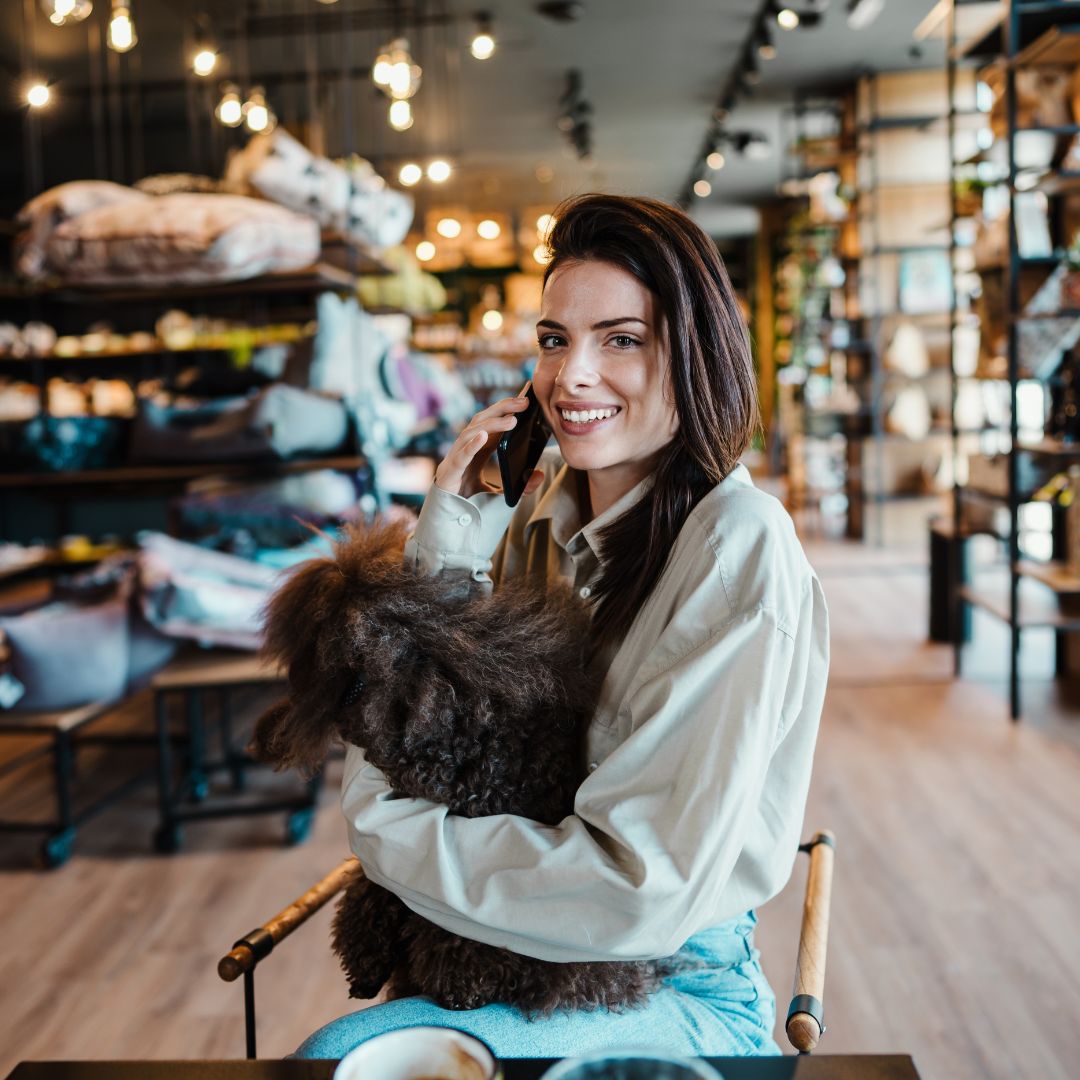 A stylish woman sitting in a trendy pet boutique, holding a curly-haired dog in her arms while talking on the phone and smiling. Shelves filled with pet supplies and decorative lights are visible in the background.