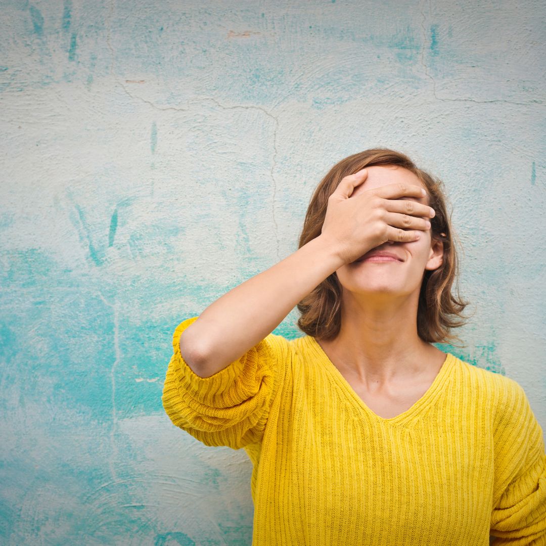 A young woman wearing a bright yellow sweater playfully covering her face with one hand, smiling with a slightly amused expression against a textured blue-green background.