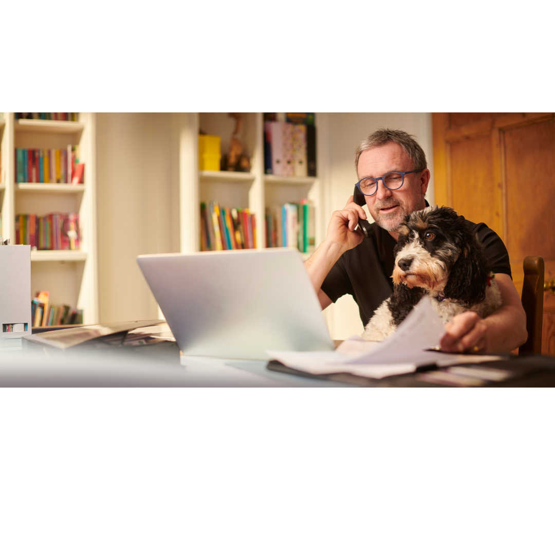A man works on his laptop while on the phone, with his dog sitting on his lap. A cosy home office with bookshelves is in the background.