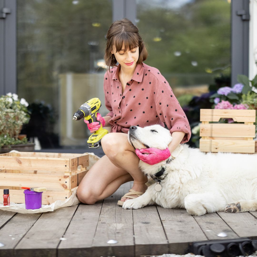 Woman holding a drill while petting a large white dog on a garden deck, surrounded by crates and plants, symbolising hands-on, DIY-style pet marketing.