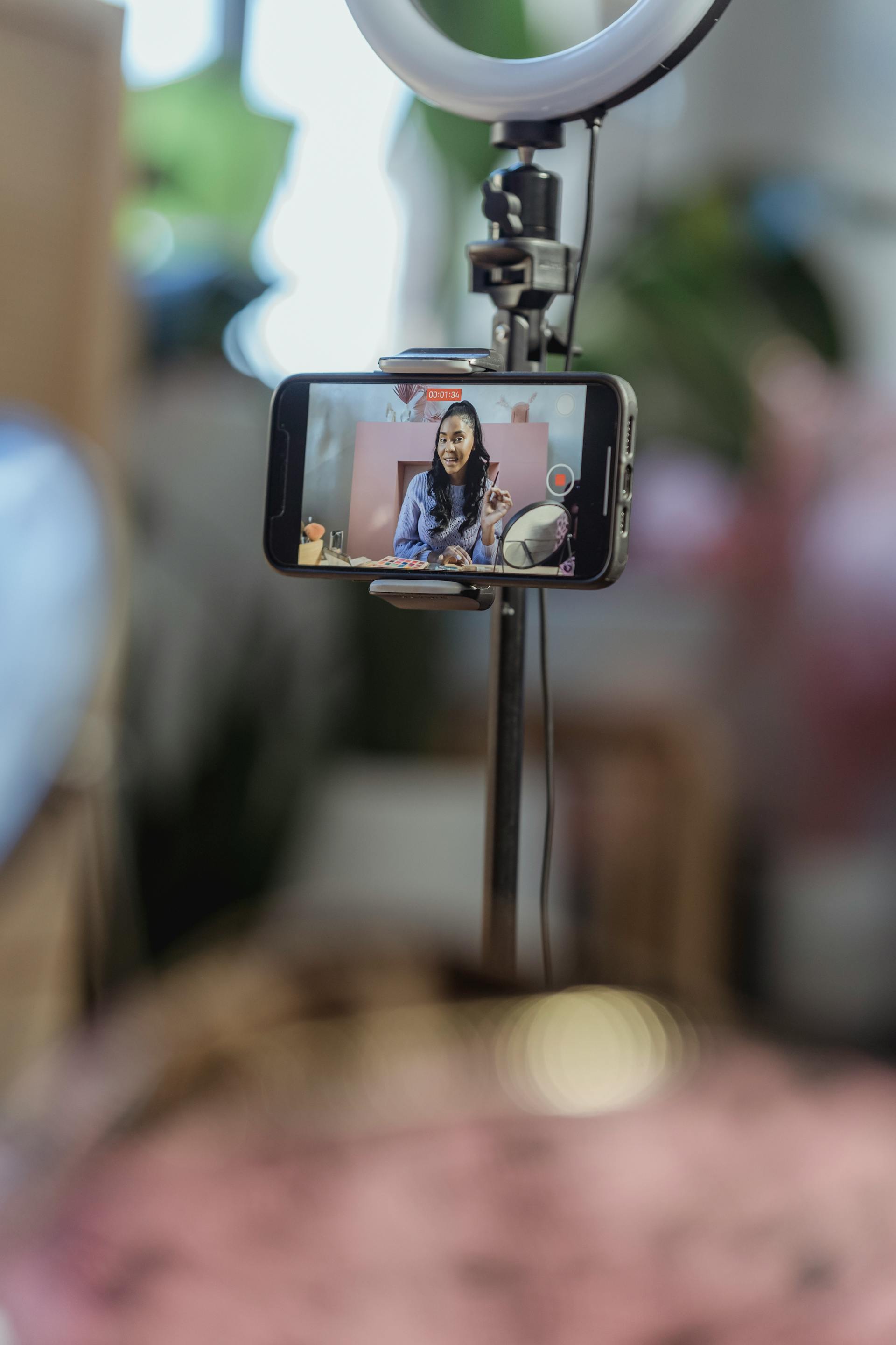 A smartphone mounted on a tripod with a ring light, capturing a woman live streaming beauty or lifestyle content.