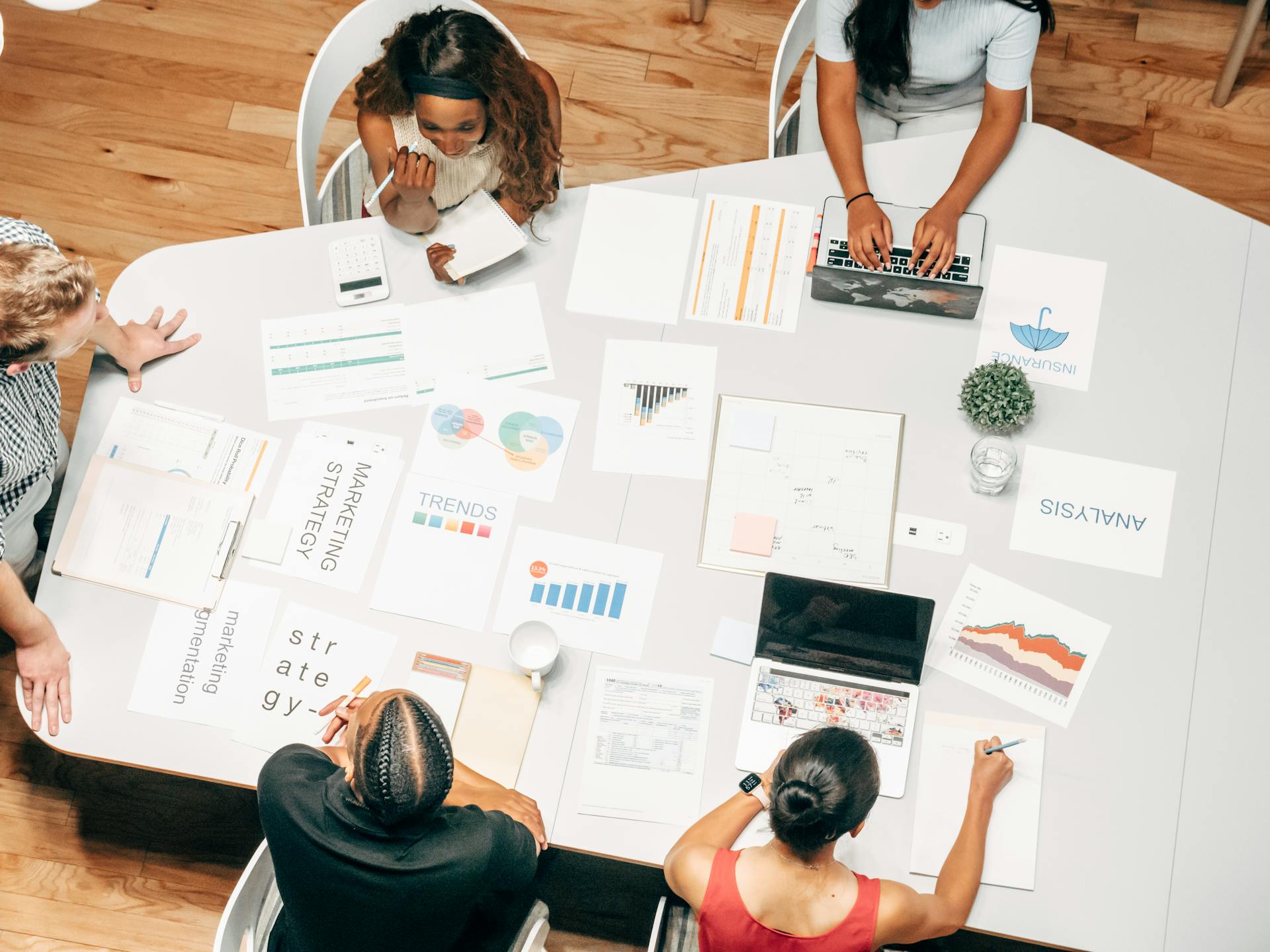 A diverse group of professionals gathered around a table, discussing marketing strategies with charts, reports, and laptops spread across the table.