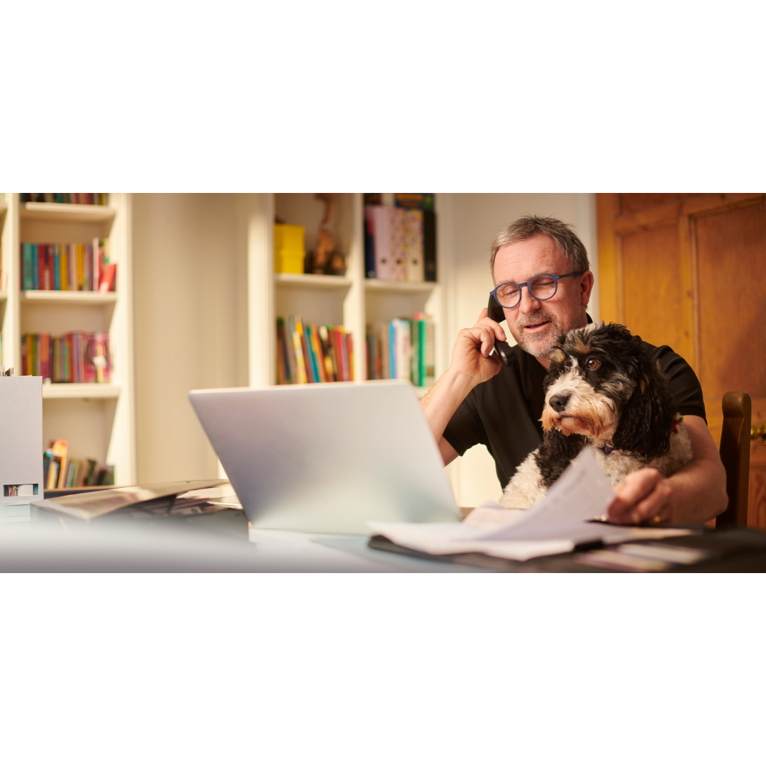 Middle aged man working from home with a laptop and phone, holding papers while his dog sits on his lap in a cosy home office.