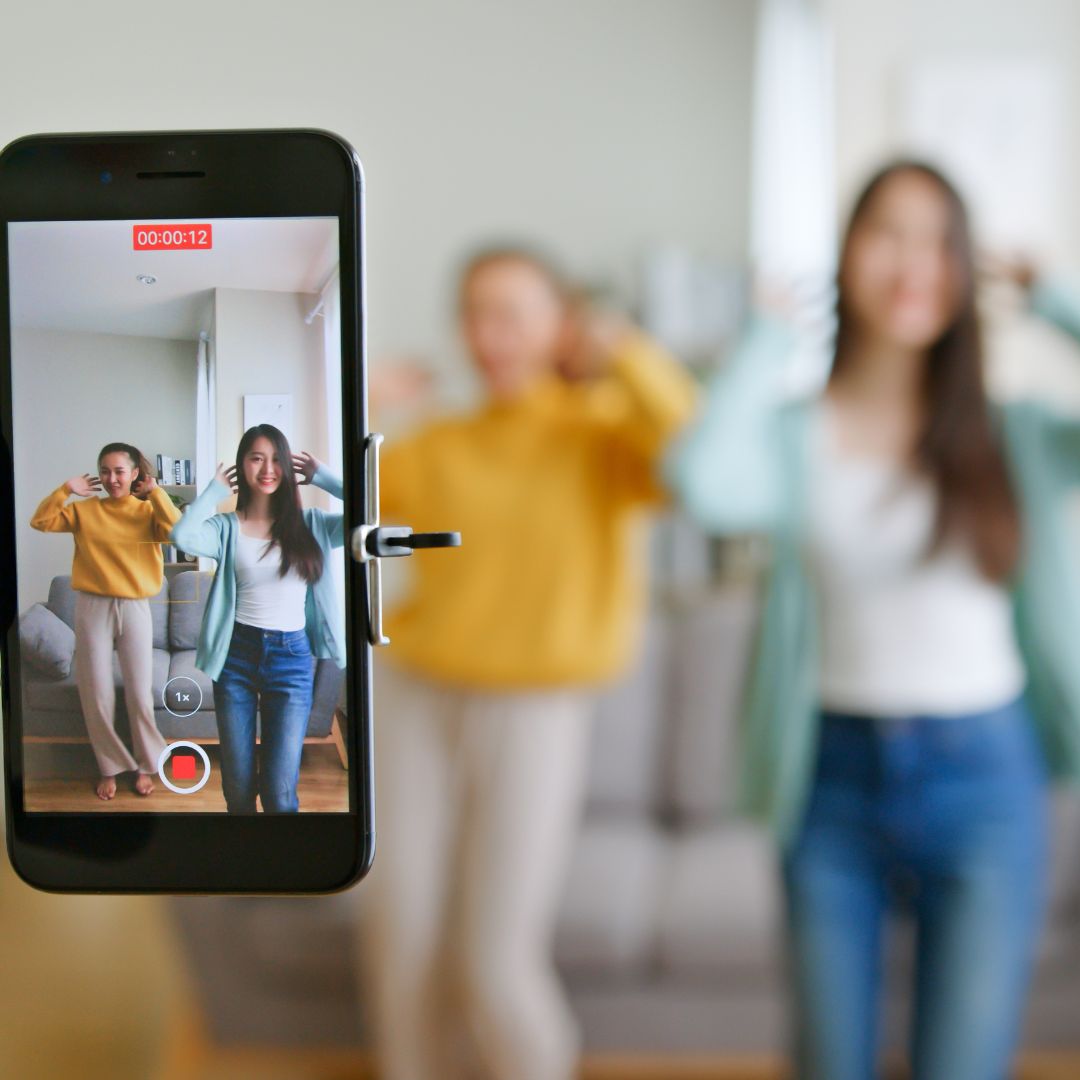 Two women record a TikTok-style dance video in a living room using a phone on a tripod.
