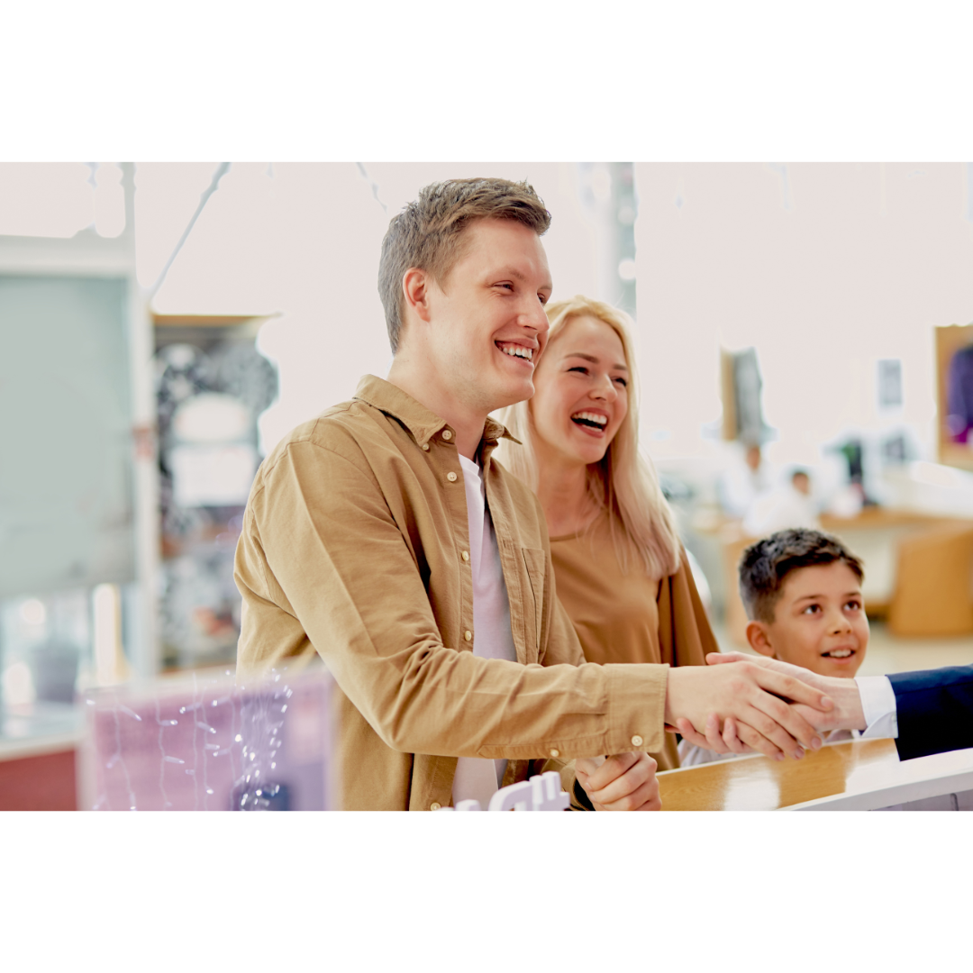 Smiling family with a young child shaking hands with a shopkeeper at a bright and welcoming retail counter.