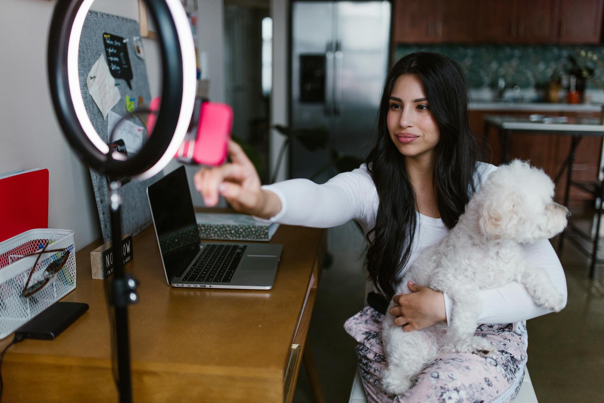 A woman sitting at a desk in her home, adjusting a smartphone mounted on a tripod with a ring light while holding a fluffy white dog in her lap. A laptop and other office items are on the desk.