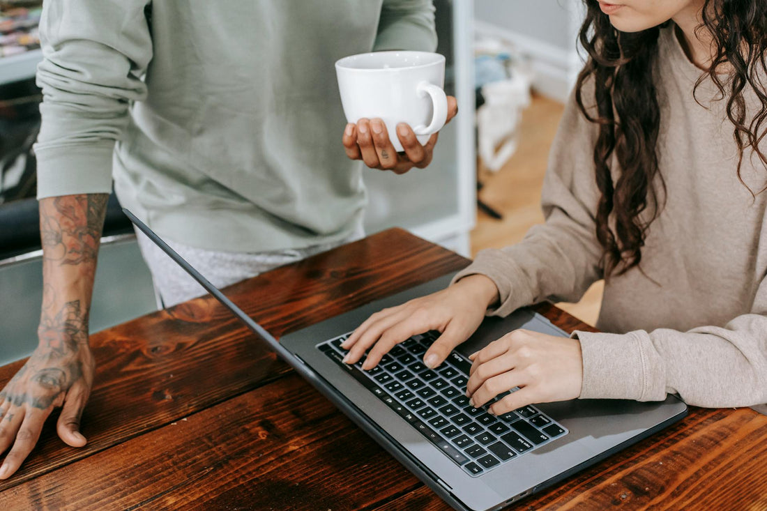 A person typing on a laptop at a rustic wooden table, while another person, holding a coffee mug, stands nearby. Their casual clothing and tattoos suggest a relaxed, creative workspace.