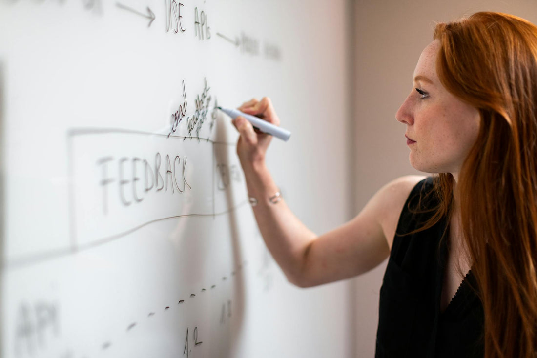 A woman with red hair writing on a whiteboard filled with diagrams and notes, actively engaged in a brainstorming or planning session.